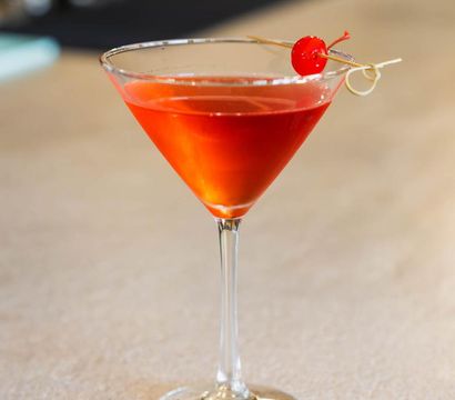 Vibrant red cocktail in a martini glass topped with a maraschino cherry on a cocktail pick, set on a light bar counter.