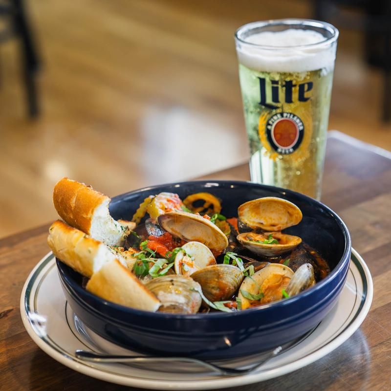 Bowl of steamed clams in a tomato broth with fresh herbs, toasted baguette slices on the rim, and a tall glass of beer on a wooden table