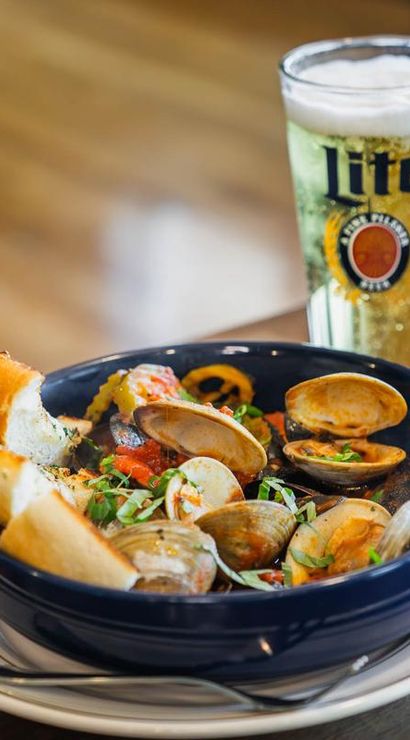 Bowl of steamed clams in a tomato broth with fresh herbs, toasted baguette slices on the rim, and a tall glass of beer on a wooden table