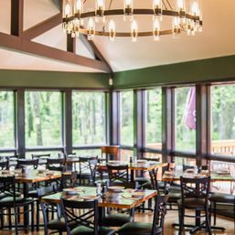 Cozy, airy restaurant dining room with wooden tables and black chairs set for service, large windows revealing a green forest and outdoor deck, and a circular chandelier hanging from a vaulted ceiling.