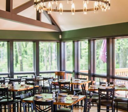 Cozy, airy restaurant dining room with wooden tables and black chairs set for service, large windows revealing a green forest and outdoor deck, and a circular chandelier hanging from a vaulted ceiling.
