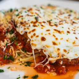 Close-up of Italian-American chicken parmesan: breaded cutlet topped with bubbly browned mozzarella over spaghetti and marinara sauce, sprinkled with chopped parsley.
