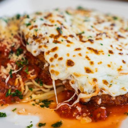 Close-up of Italian-American chicken parmesan: breaded cutlet topped with bubbly browned mozzarella over spaghetti and marinara sauce, sprinkled with chopped parsley.
