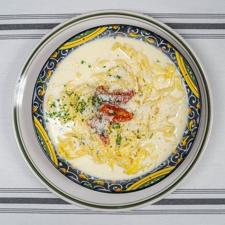 Creamy fettuccine Alfredo with grated Parmesan, chopped parsley and sun-dried tomatoes in a decorative Mediterranean-style bowl on a striped tablecloth