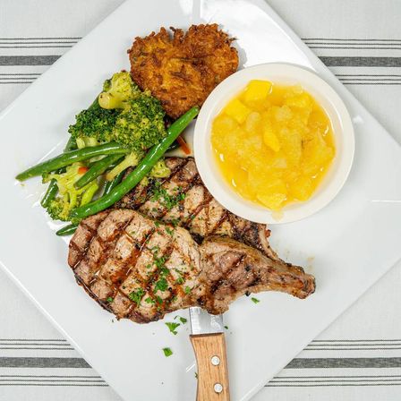 Hearty American grilled bone-in pork chop with char marks, sautéed green beans and broccoli, crispy potato pancake, and a bowl of sweet peach compote on a white plate.