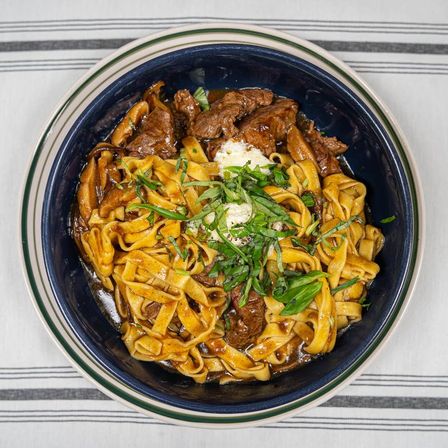 Top-down view of hearty pappardelle pasta with braised beef ragu, a dollop of ricotta and sliced basil in a blue bowl on a striped tablecloth.