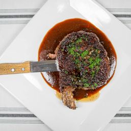 Overhead close-up of a seared bone-in ribeye on a white square plate, resting in rich brown sauce, garnished with chopped parsley and served with a wooden-handled steak knife.
