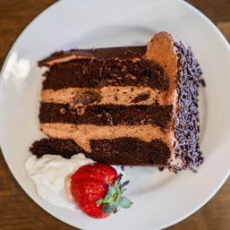Three-layer chocolate cake slice with chocolate frosting and sprinkles on a white plate, served with whipped cream and a fresh strawberry.