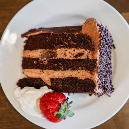 Three-layer chocolate cake slice with chocolate frosting and sprinkles on a white plate, served with whipped cream and a fresh strawberry.