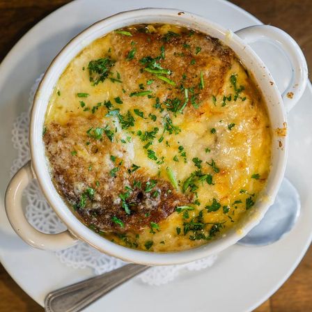 Bistro-style French onion soup in a white handled bowl, bubbling golden melted cheese over toasted bread, sprinkled with chopped parsley and served with a spoon on a plate.