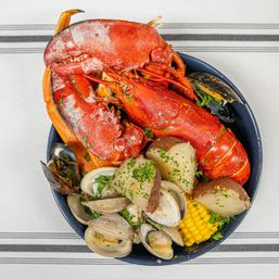 Top-down view of a New England-style seafood boil: bright red whole lobster, clams and mussels, boiled red potatoes, corn on the cob, and chopped parsley in a bowl on a striped tablecloth