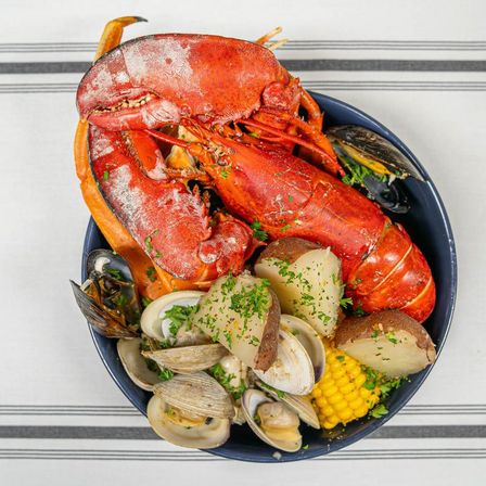 Top-down view of a New England-style seafood boil: bright red whole lobster, clams and mussels, boiled red potatoes, corn on the cob, and chopped parsley in a bowl on a striped tablecloth