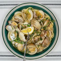 Top-down view of clam linguine: spaghetti with steamed clams in a garlicky white-wine broth, lemon wedge and chopped parsley in a green-rimmed bowl on a striped tablecloth.