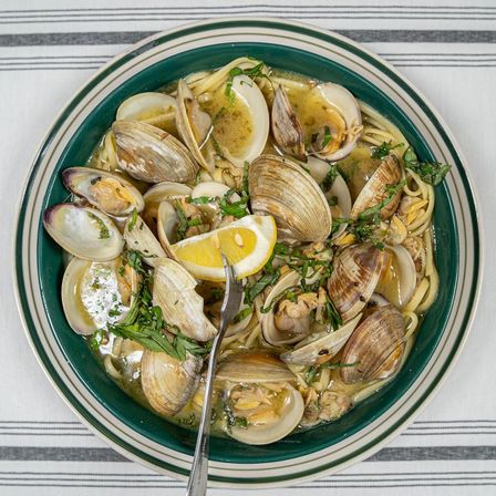 Top-down view of clam linguine: spaghetti with steamed clams in a garlicky white-wine broth, lemon wedge and chopped parsley in a green-rimmed bowl on a striped tablecloth.
