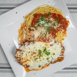 Top-down shot of classic chicken Parmesan—crispy breaded cutlet with melted mozzarella and parsley—served with spaghetti marinara and grated Parmesan on a white plate.