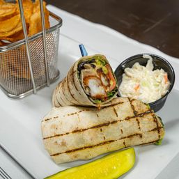 Grilled shrimp and avocado wrap, halved to show shrimp, lettuce and tomato, served on a white plate with coleslaw in a black cup, a metal basket of chips and a dill pickle spear.