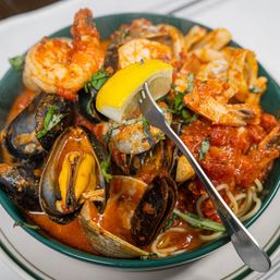 Close-up of Mediterranean-style seafood pasta in a green bowl: mussels, clams and shrimp over spaghetti in spicy tomato sauce, garnished with fresh basil and a lemon wedge on a fork.