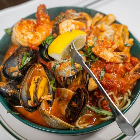 Close-up of Mediterranean-style seafood pasta in a green bowl: mussels, clams and shrimp over spaghetti in spicy tomato sauce, garnished with fresh basil and a lemon wedge on a fork.