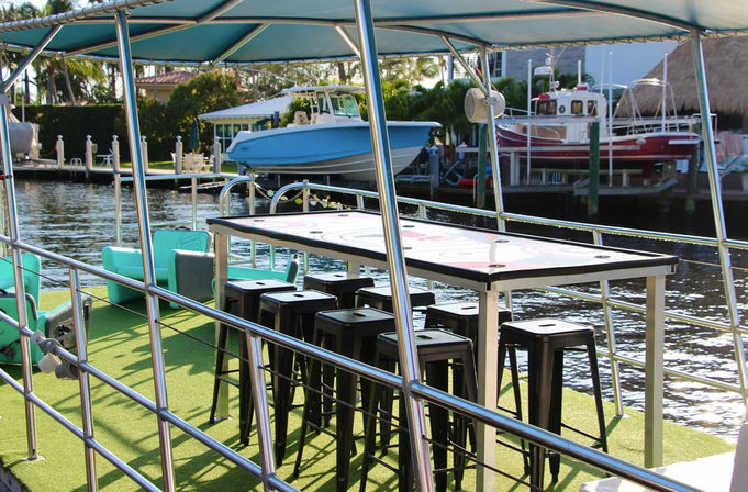 Sunlit boat deck with a bar-height table and black stools under a blue canopy on green turf, overlooking a canal marina with docked boats and docks.