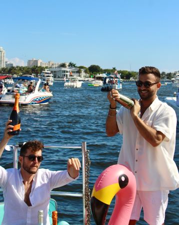 Boat party on a sunny waterway with two men celebrating—one shaking a cocktail shaker, the other holding a champagne bottle, a pink inflatable flamingo in the foreground and boats and waterfront skyline in the background.