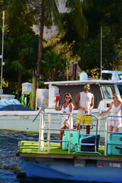 Three people on a turquoise pontoon with green turf at a sunny Florida marina — woman raising a red cup, man in yellow shorts and sunglasses, another woman in white, palm trees and motorboats in the background.