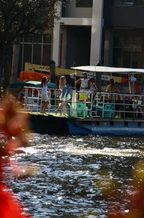 Group of people enjoying a sunny pontoon boat party along an urban riverfront, colorful seating and sparkling water