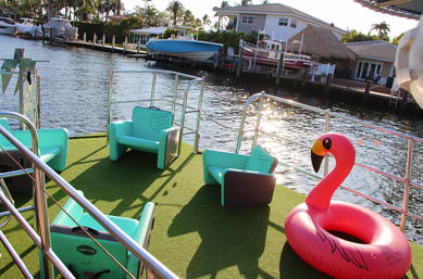 Sunlit waterfront canal deck with turquoise lounge chairs on green turf, a bright pink inflatable flamingo float, stainless railings, and boats and palm trees along the dock
