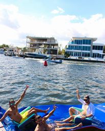 Three men relaxing and waving on a bright blue inflatable raft in a sunny waterfront canal, with motorboats and modern luxury waterfront homes and docks along the shoreline.