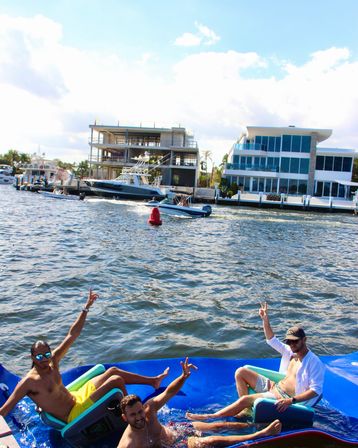 Three men relaxing and waving on a bright blue inflatable raft in a sunny waterfront canal, with motorboats and modern luxury waterfront homes and docks along the shoreline.