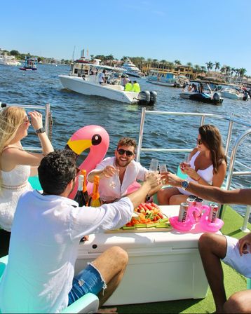 Friends toasting with champagne on a sunny boat party at a palm-lined marina — inflatable pink flamingo, fruit platter and drinks on deck, motorboats and waterfront homes in the background.