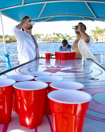 Boat party beer pong: red solo cups lined up on a table aboard a sunny coastal boat, two shirted men chugging drinks with palm-tree shoreline in the background.