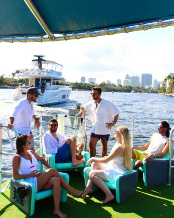 Group of friends relaxing on a sunlit party boat in South Florida waterways, turquoise deck chairs with a luxury yacht and city skyline in the background.