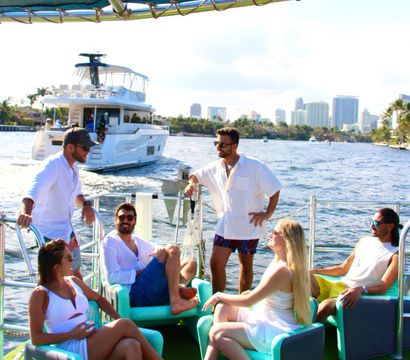 Group of friends relaxing on a sunlit party boat in South Florida waterways, turquoise deck chairs with a luxury yacht and city skyline in the background.