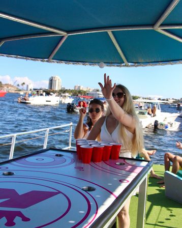 Friends playing beer pong on a sunny party boat — ping-pong ball midair above red cups on the table, with boats and a coastal marina skyline in the background.