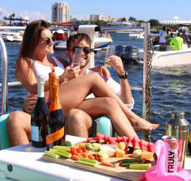 Couple toasting with champagne on a sunny boat party at a busy marina, picnic board of fruit, veggies and bottles in the foreground