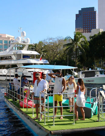 Friends enjoying a sunny day on a turquoise party pontoon boat in a busy marina with luxury yachts, palm trees, and downtown waterfront high-rises in the background.