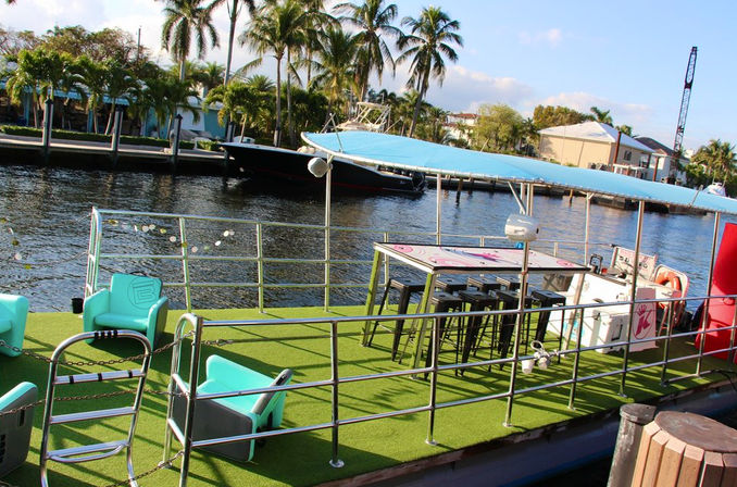 Waterfront party boat docked on a tropical canal with palm trees, artificial turf deck, turquoise lounge chairs, bar seating and blue canopy.