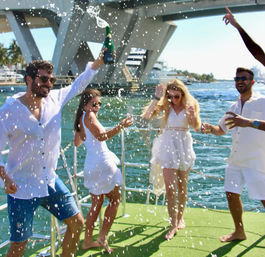 Group of four friends in white popping champagne and dancing on a sunny boat deck with yachts, a bridge, and marina in the background