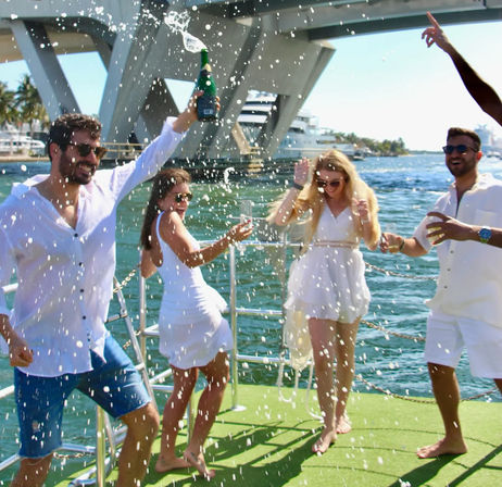 Group of four friends in white popping champagne and dancing on a sunny boat deck with yachts, a bridge, and marina in the background