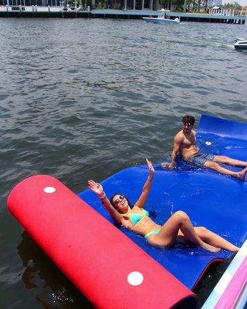 Young adults enjoying summer at a marina: woman in a turquoise bikini laughing with arms raised and man reclining on a blue inflatable floating mat beside a red buoy, boats and waterfront buildings in the background.