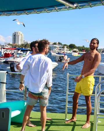 Three friends laughing on a boat platform at a busy marina, one in yellow swim trunks, seagulls overhead and boats and waterfront buildings under a clear blue sky
