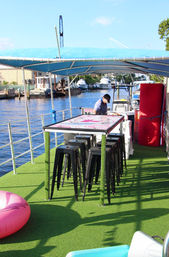 Sunny waterfront boat deck at a marina with blue canopy, green artificial turf, a tall table surrounded by black bar stools, a pink inflatable tube and a person at the helm with yachts docked in the background.