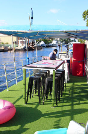 Sunny waterfront boat deck at a marina with blue canopy, green artificial turf, a tall table surrounded by black bar stools, a pink inflatable tube and a person at the helm with yachts docked in the background.