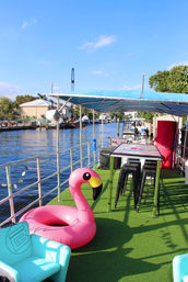 Sunny waterfront canal boat deck with green turf, party-ready pink inflatable flamingo float, high bar table and black stools under a blue canopy, with boats and waterfront homes in the background.