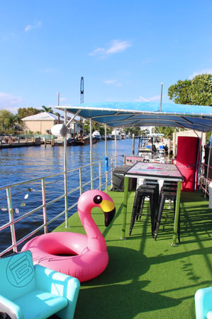 Sunny waterfront canal boat deck with green turf, party-ready pink inflatable flamingo float, high bar table and black stools under a blue canopy, with boats and waterfront homes in the background.