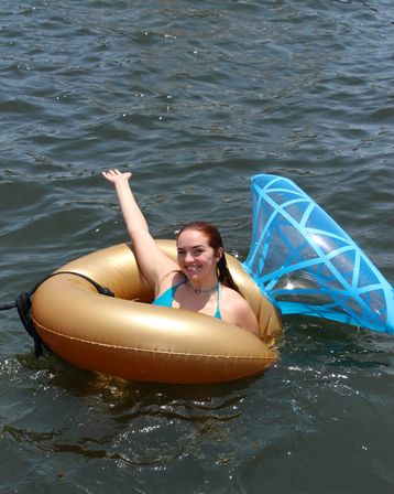 Smiling woman in a teal bikini floating on a gold inner tube with a blue geometric float in open water, waving during a sunny summer outing.