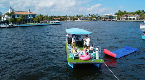 Party pontoon boat with blue canopy, people and pink flamingo float near luxury waterfront homes along a sunny coastal waterway.