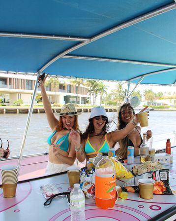 Three friends in colorful bikinis cheering with plastic cups on a sunny boat party under a blue canopy, snacks and drinks on the table and waterfront homes along a canal in the background.