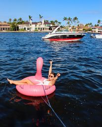 Person relaxing on a bright pink flamingo float in deep blue water, tethered by a rope, smiling and pointing toward a red-and-black powerboat with luxury waterfront homes and palm trees lining the sunny shoreline.