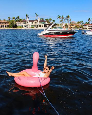 Person relaxing on a bright pink flamingo float in deep blue water, tethered by a rope, smiling and pointing toward a red-and-black powerboat with luxury waterfront homes and palm trees lining the sunny shoreline.